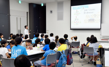 ワークショップ中の様子。BIKEN職員がモニターを使って説明をしており、大勢の小学生がそれを聞いている。